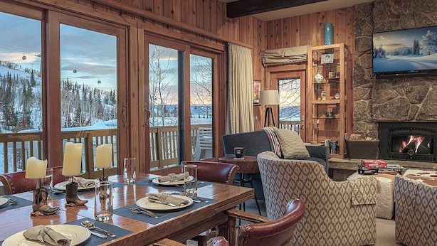 A cozy cabin interior with a dining area, a fireplace, and large windows overlooking a snowy landscape. - Luxury + Pool/Hot Tub + Ski in/Ski out - Right-O-Way Chalet (Steamboat Springs)