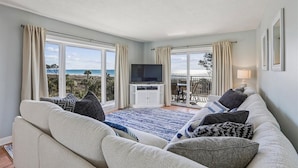 Een woonkamer met een groot raam met uitzicht op het strand, een flatscreen televisie en een balkon met een tafel en stoelen.