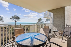 Een balkon met een ronde tafel en stoelen, met uitzicht op een strand met bomen en een heldere lucht.