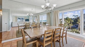 Een ruime eetruimte met een grote houten tafel en stoelen, een kroonluchter en grote ramen met uitzicht op de omgeving.