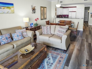 Una sala de estar con sofá, mesa de centro y alfombra. Se ve una cocina al fondo con armarios blancos y un comedor.