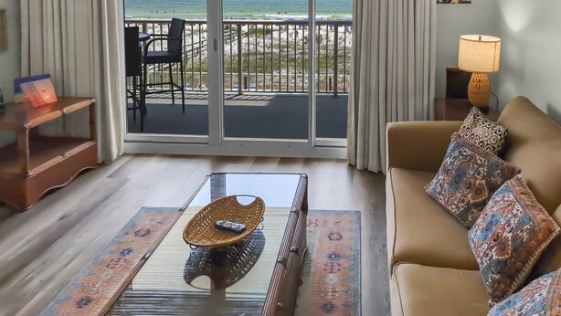 A hotel room with a balcony overlooking a beach, a sofa with patterned cushions, a woven coffee table with a remote control, and a lamp on a side table.