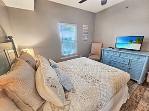 Een slaapkamer met een bed, een stoel, een kaptafel, een televisietoestel en een raam met jaloezieën.