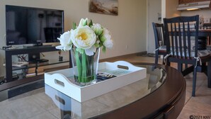 Ein Wohnzimmer mit einem Glastisch, einer Vase mit weißen Blumen, einem Flachbildfernseher und einem Gemälde an der Wand.