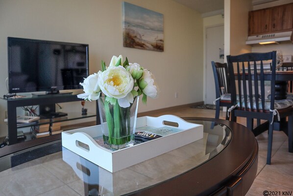 Un salon avec une table basse en verre, un vase rempli de fleurs blanches, un téléviseur à écran plat et un tableau accroché au mur.