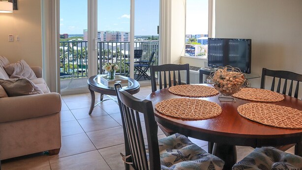 Ein Wohnzimmer mit einer Couch, einem Esstisch mit Platzsets, einem Fernseher und einem Balkon mit Blick auf Gebäude und Grünflächen.