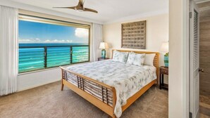 A bedroom with a large window offering a view of the ocean, a bed with a wooden frame, a ceiling fan, and a nightstand with a lamp.