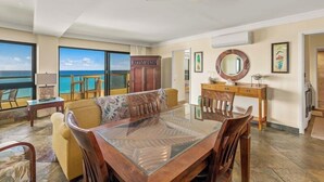 A spacious living area with a large glass table, wooden chairs, a sofa, and a ceiling fan. A view of the ocean is visible through large windows.