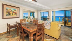 A spacious living room with a wooden dining table, yellow sofa, and large windows offering ocean views.
