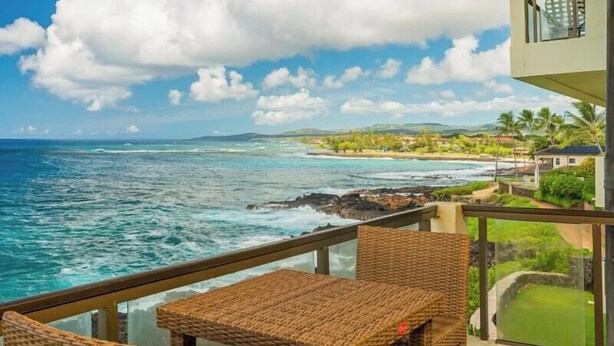 A balcony with wicker furniture overlooking a coastal view with the ocean and a distant shoreline with buildings and greenery.