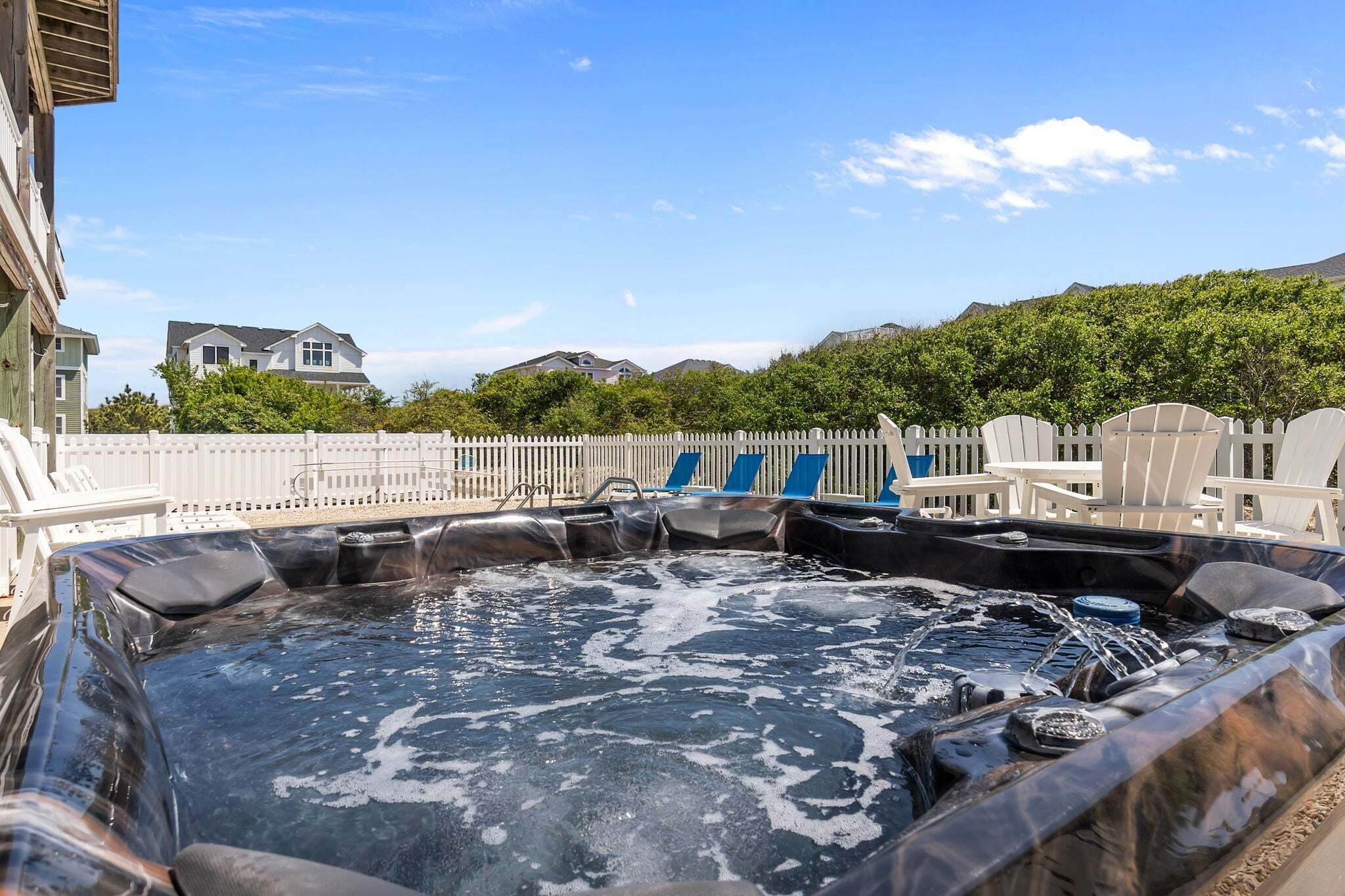 A hot tub with blue chairs, a white fence, and houses in the background.
