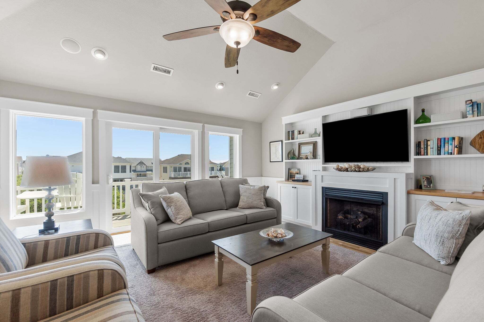 A spacious living room with a fireplace, a ceiling fan, and large windows.