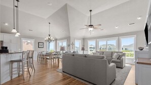 A spacious living area with a large sofa, dining table, and chairs, a kitchen with white cabinets, and large windows offering views of the outside. - Only 70 Steps to beach access! Elevator! Heated Pool!  Pets, Pool Table, Hot tub (Corolla)