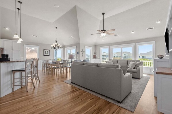 A spacious living area with a large sofa, dining table, and chairs, a kitchen with white cabinets, and large windows offering views of the outside. - Only 70 Steps to beach access! Elevator! Heated Pool!  Pets, Pool Table, Hot tub (Corolla)