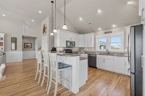 A modern kitchen with white cabinets, a central island with bar stools, a stainless steel refrigerator, and a view of the outdoors through large windows. - Only 70 Steps to beach access! Elevator! Heated Pool!  Pets, Pool Table, Hot tub (Corolla)