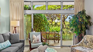 Un salón con un sofá gris, un sillón estampado y una mesa de centro. Hay un ventanal con cortinas, una planta y vistas a la vegetación exterior.