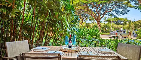 An outdoor dining area with a wooden table set for four, surrounded by lush greenery and a view of a road with a vehicle.