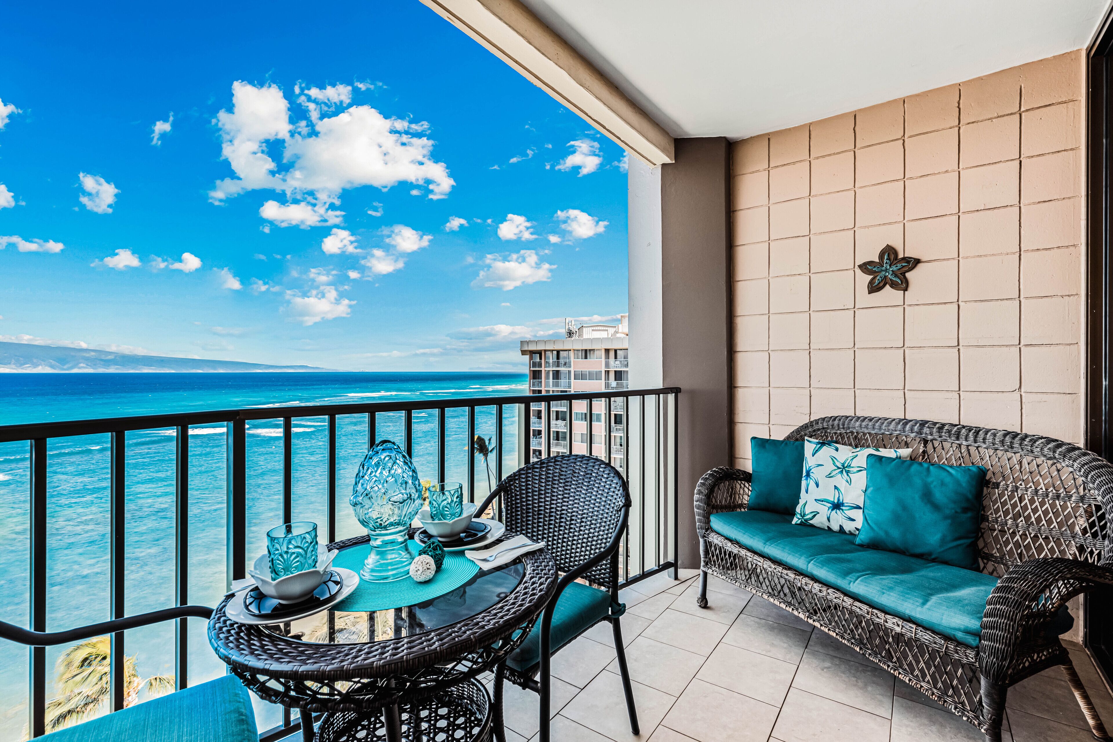 Un balcon avec vue sur l’océan, un canapé en osier avec des coussins turquoise et une table à manger avec de la verrerie.