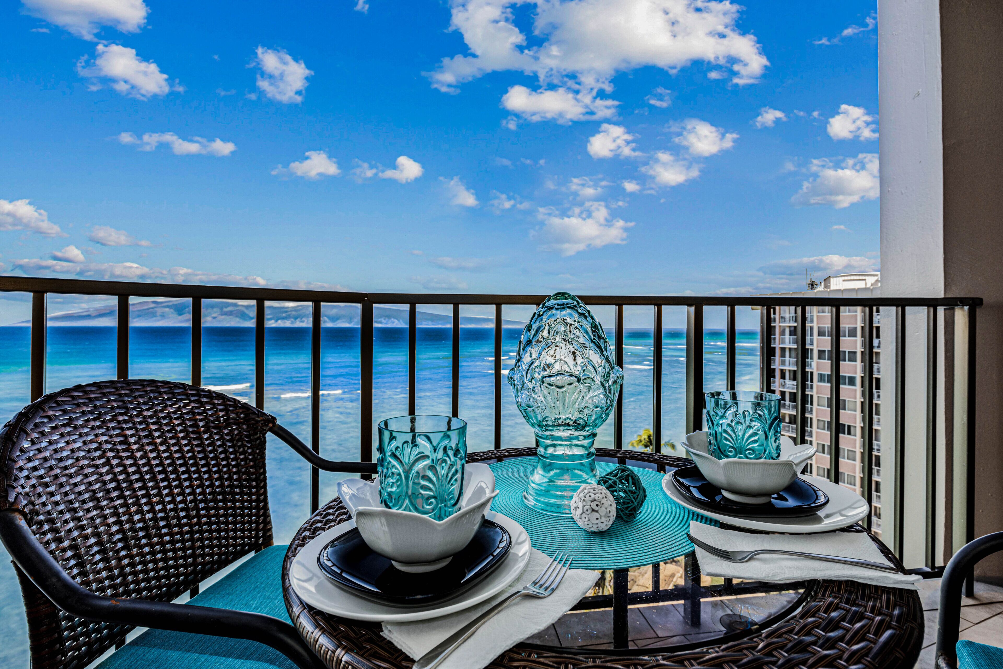 A balcony with a view of the ocean, two wicker chairs, a table set with glassware, and a railing.