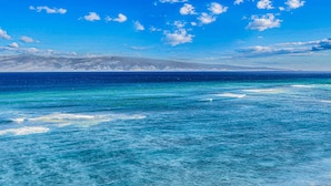 Une vue dégagée sur un paysage côtier, avec une mer calme et des montagnes lointaines, le tout sous un ciel bleu parsemé de nuages.