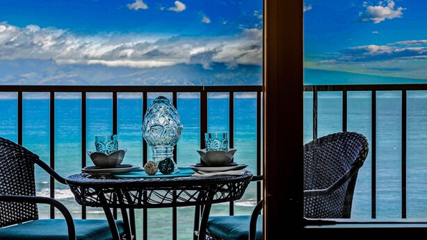 Un balcon avec une table dressée pour deux personnes, offrant une vue imprenable sur une mer et un ciel d’un bleu éclatant.