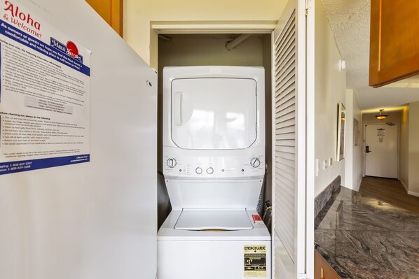 A laundry room with a front-loading washer and dryer, a bulletin board with a welcome sign, and a hallway leading to another room.