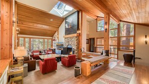 Amplia sala de estar con chimenea de piedra, techo de madera y ventanas grandes que ofrecen vistas al exterior.