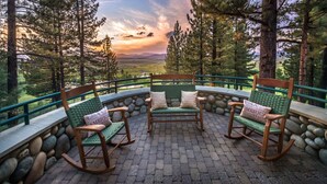 Un patio con sillas mecedoras y un muro de piedra, con vistas a un paisaje pintoresco con árboles y una puesta de sol.
