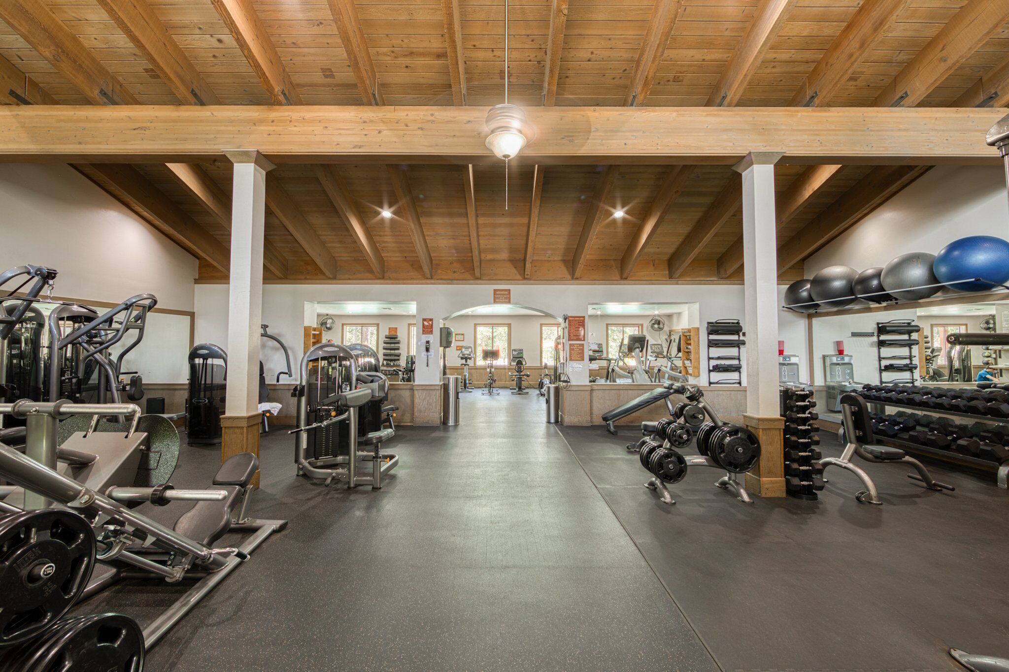 Un gimnasio con varios equipos de ejercicio, vigas de madera en el techo y un acceso central.