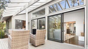 Une maison moderne dotée d’une terrasse, de fauteuils en osier et d’une grande porte-fenêtre coulissante qui donne sur un intérieur comprenant un canapé et une lampe.