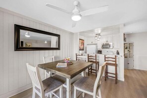 A dining area with a wooden table and white chairs, a kitchen with white cabinets, and a ceiling fan.