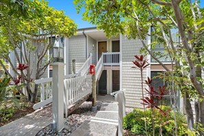 A residential building with a staircase leading to a second-floor balcony, surrounded by greenery and a white fence. - Ground floor condo with grassy area, quick Beach Access, G33 North Coast Village (Oceanside)