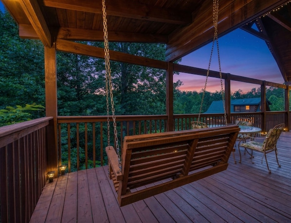 A wooden deck with a swing, a table with chairs, and a view of trees and a building in the background.