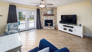 A living room with a stone fireplace, a flat-screen TV, a ceiling fan, and a large window with curtains. - 356 Billfish🐟Beachside Townhome🐶Pet Friendly (Fort Walton Beach)