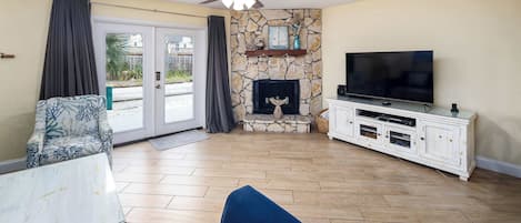 Una sala de estar con chimenea de piedra, televisor de pantalla plana, ventilador de techo y un ventanal con cortinas.