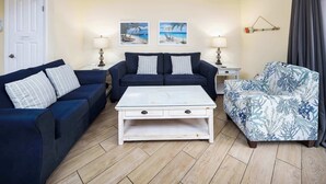 Un soggiorno con un divano blu scuro, un tavolino da caffè bianco e una poltrona con motivi floreali. Si notano due lampade a parete e due quadri con scene di spiaggia.
