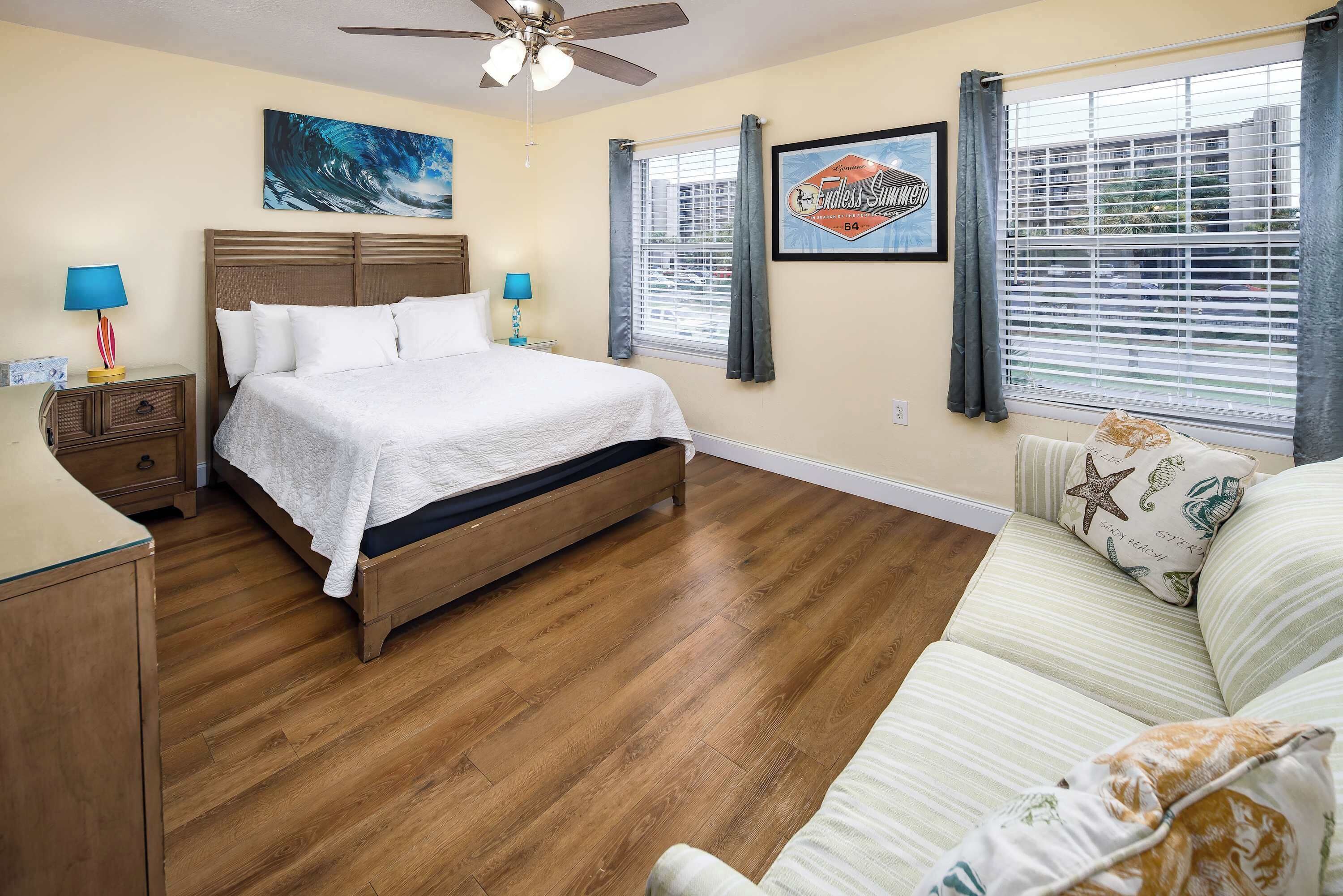 A bedroom with a wooden bed, a nightstand with a lamp, a window with blinds, and a wall with a framed picture.