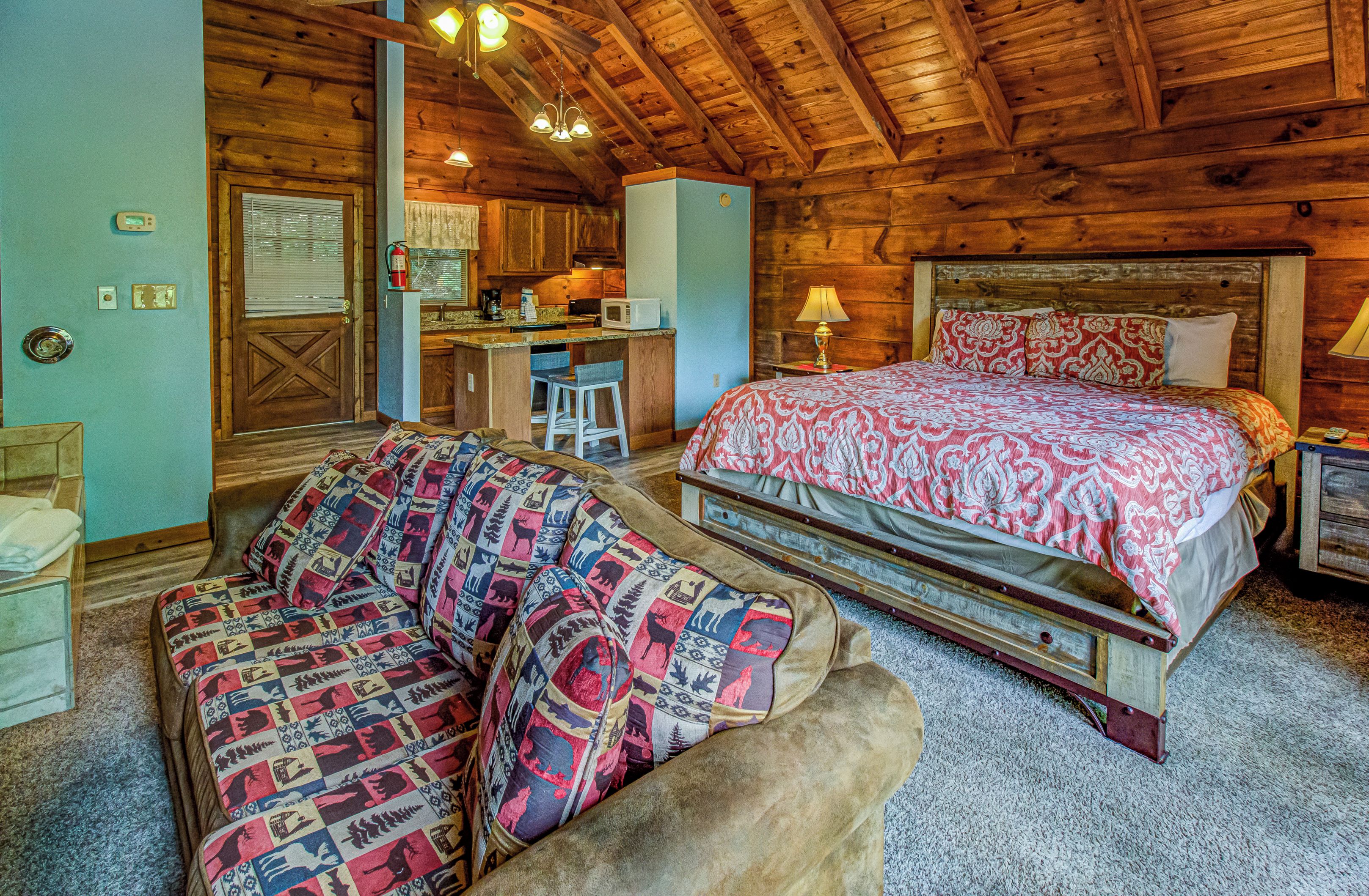 A cozy log cabin room with a bed, a sofa, a kitchenette, and a bathroom.