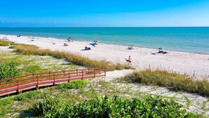 Ein Strand mit einer Holzpromenade, die zum Ufer führt, klarem blauen Wasser und Menschen, die am Sand entspannen.