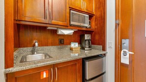 Una cocina compacta con armarios de madera, un lavamanos de acero inoxidable, un microondas, una cafetera y un refrigerador.