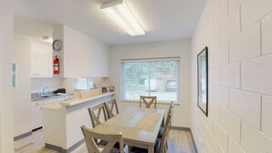 Una cocina compacta con un lavamanos, encimera y un reloj en la pared. Hay una mesa de comedor con sillas y una ventana con persianas.