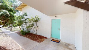Un edificio de ladrillo blanco con una puerta azul turquesa, un árbol pequeño y un sendero de hormigón.