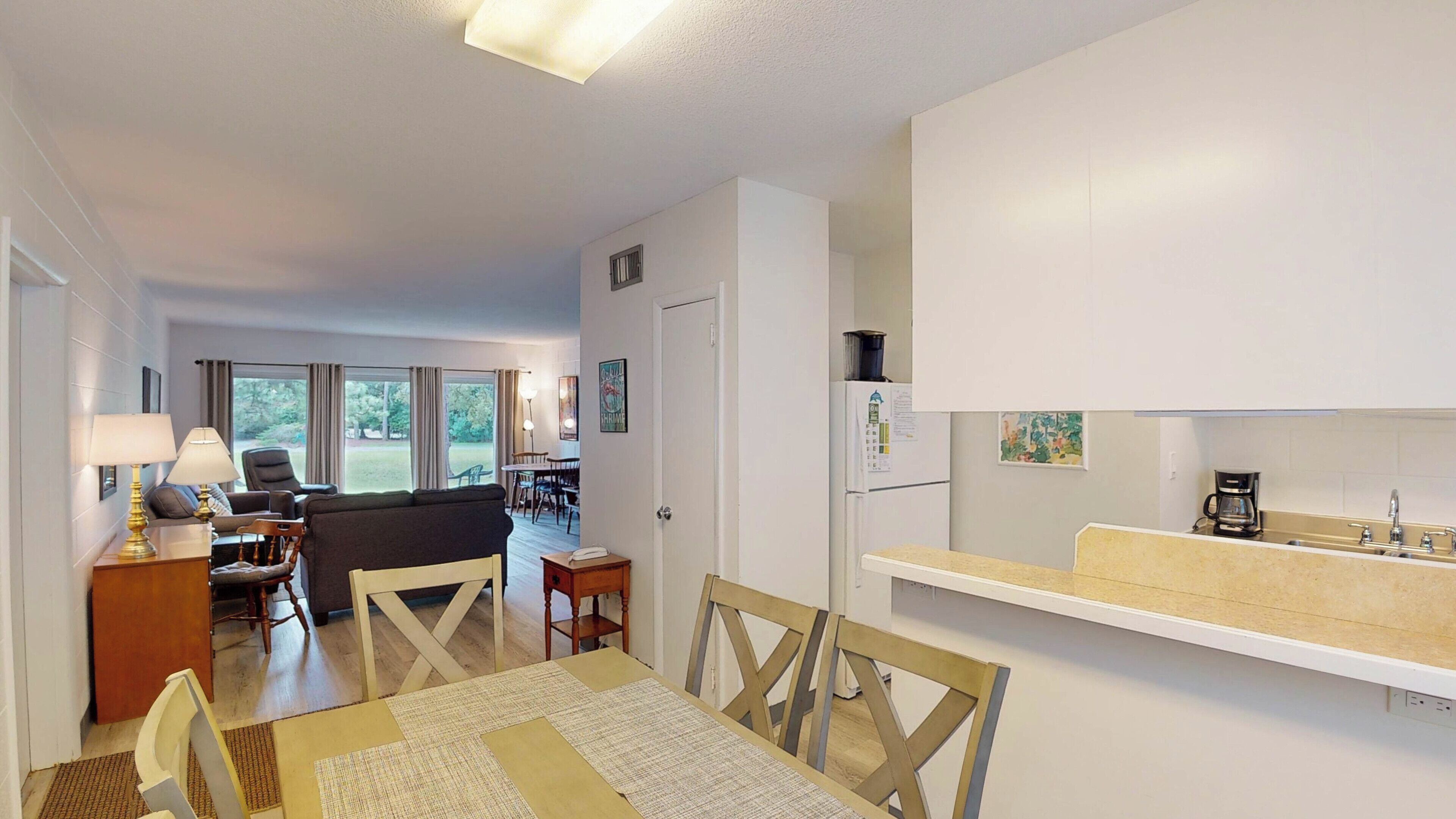 Una cocina moderna con armarios blancos, cafetera y lavamanos. Hay una zona de comedor con mesa y sillas.