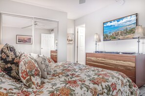 Un dormitorio con colcha estampada, cómoda de madera, televisor de pantalla plana y ventilador de techo.