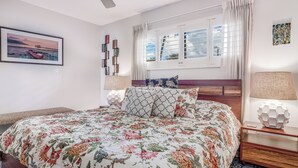A neatly arranged bedroom with a large bed, a window with curtains, a bedside table with a lamp, and a framed picture on the wall. - Kihei Beach #207 – Your Perfect Oceanfront Escape (Kihei)