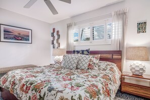A neatly arranged bedroom with a large bed, a window with curtains, a bedside table with a lamp, and a framed picture on the wall.