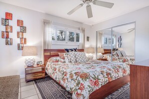 Une chambre avec un grand lit, un ventilateur de plafond, une table de chevet avec une lampe, un miroir et une fenêtre avec des rideaux.