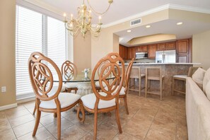 A dining area with a glass table and wooden chairs, a kitchen with wooden cabinets and stainless steel appliances, and a beige wall. - Spacious 4-bedroom beachfront end unit w/private balcony! (Destin)