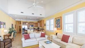 A spacious living room with a white sofa, a coffee table, and a colorful rug. There is a kitchen in the background with wooden cabinets and a dining area with wicker furniture.