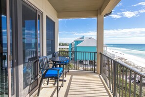 A balcony with a view of the beach, a table with chairs, and a building in the background.